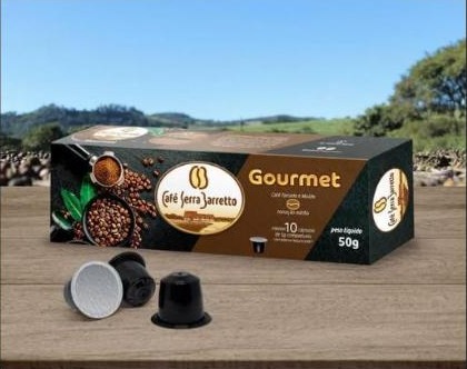 Caixa de cápsulas Gourmet sobre uma mesa de madeira, com cápsulas ao lado e o cafezal ao fundo